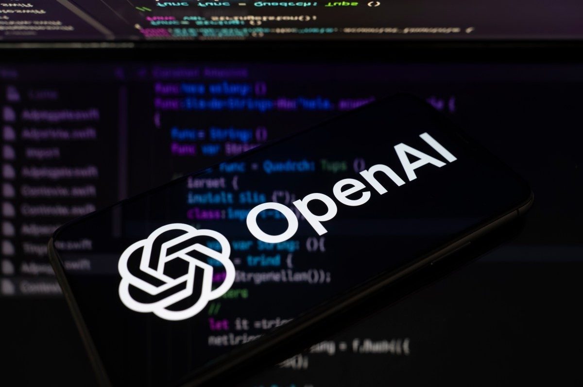 Logo da OpenAI exibido em smartphone com código ao fundo